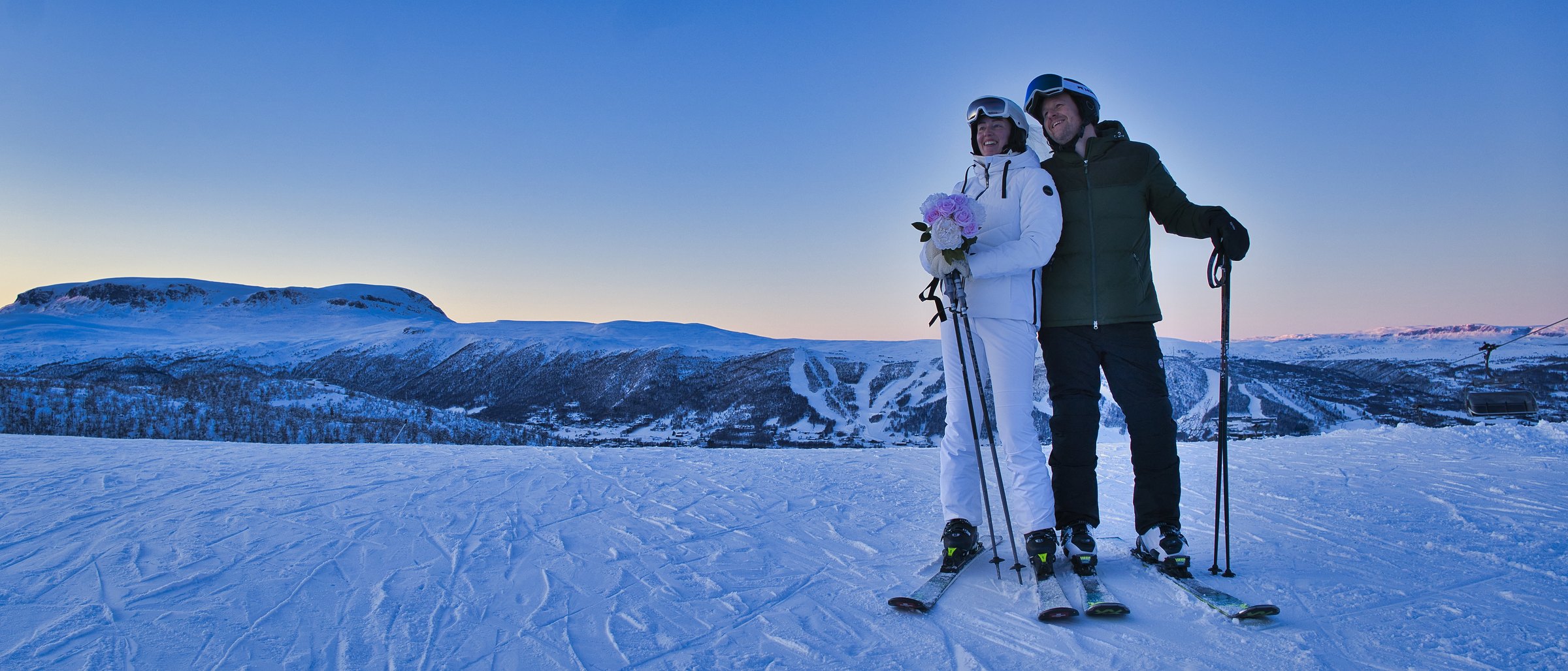 Brudepar i skiklær på et fjell ved solnedgang, med bukett og utsikt over fjellheimen