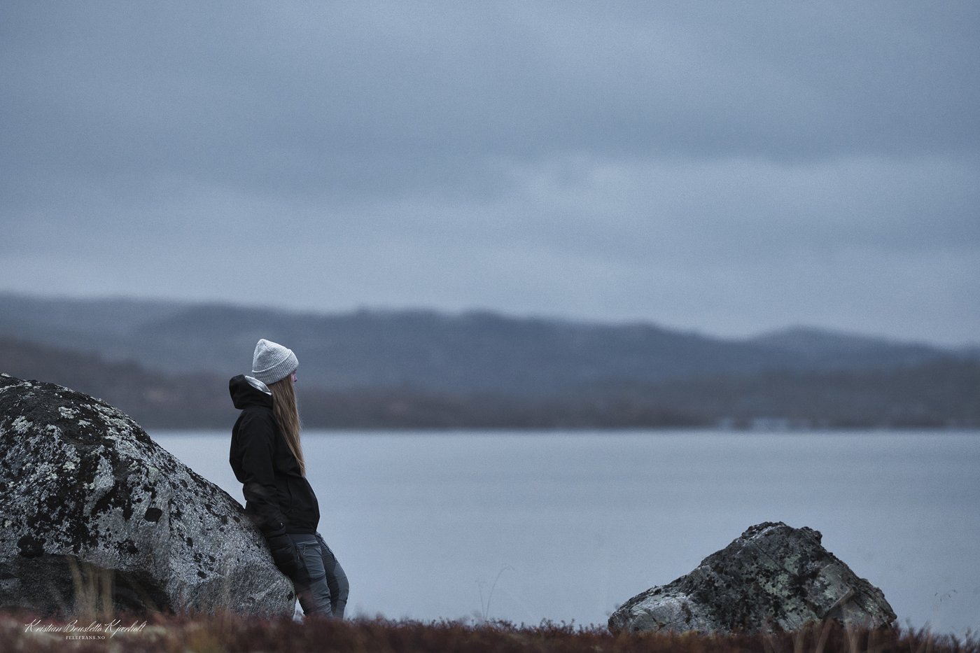 Kvinne lener seg mot en stein og ser utover et vann i disig vær